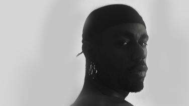 Ein Schwarzer Mann mit glänzenden Ohrringen und einem Kopftuch blickt ernst über seine Schulter in die Kamera. Schwarz-Weiß Fotografie